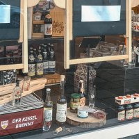 Schaufenster mit regionalen Produkten wie Bier, Spirituosen und Lebensmitteln. Dekoriert mit Pflanzen und Werbeschildern. Schriftzug 'Der Kessel brennt' sichtbar., &copy; 0711 Store, Stuttgart