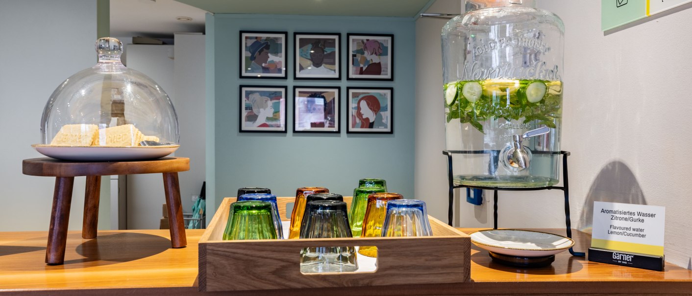 Ein Empfangstisch mit aromatisiertem Wasser, bunten Gl&auml;sern und Keksen unter einer Glasglocke. An der Wand h&auml;ngen Bilder., &copy; Novum  Hospitality