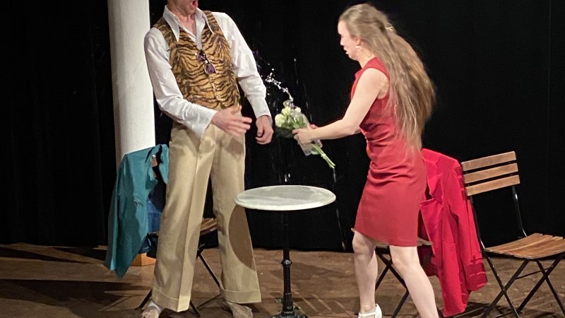 Eine Frau in rotem Kleid spritzt Wasser aus einem Blumenstrauß auf einen Mann in Tigerweste auf einer Bühne. Ein Tisch und Stühle sind im Hintergrund., © Wilhelma Theater Eine Frau in rotem Kleid spritzt Wasser aus einem Blumenstrauß auf einen Mann in Tigerweste auf einer Bühne. Ein Tisch und Stühle sind im Hintergrund., © Wilhelma Theater