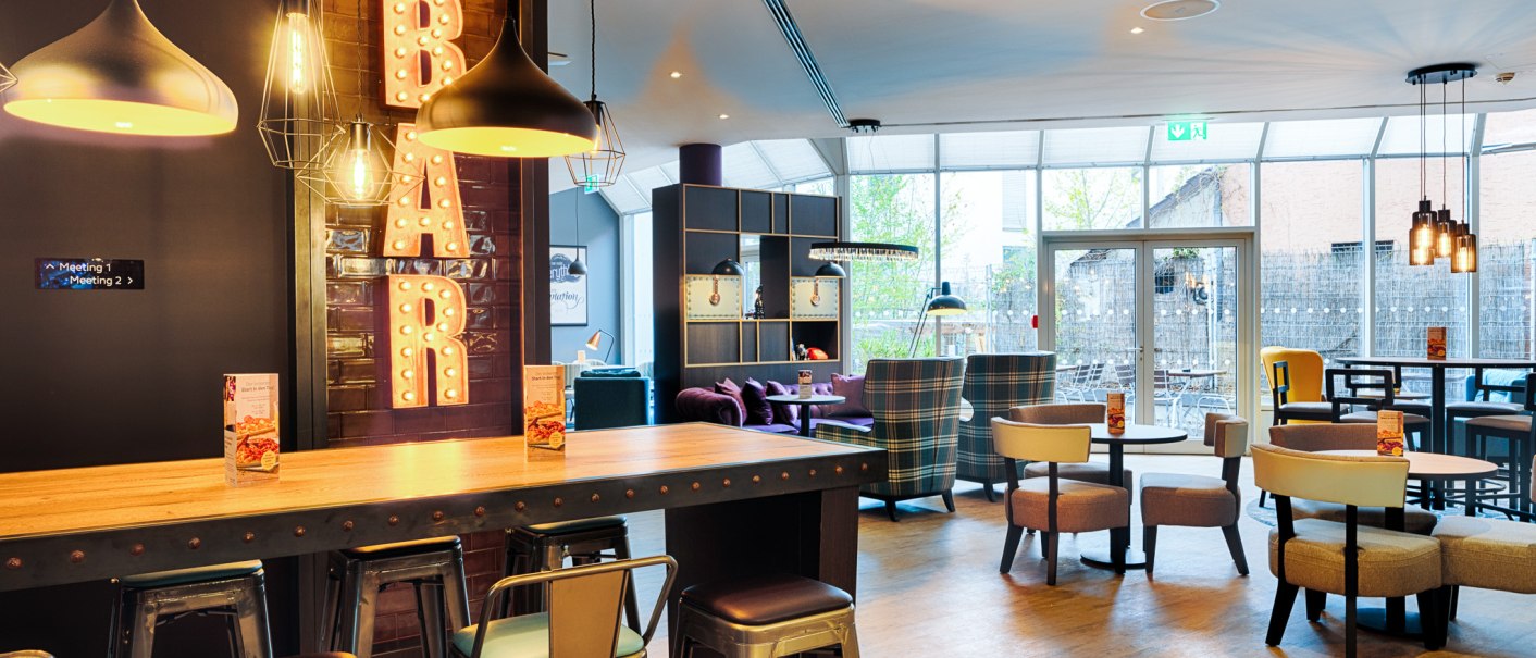 Moderne Bar mit stilvoller Beleuchtung, hohen Tischen und St&uuml;hlen. Im Hintergrund gem&uuml;tliche Sitzbereiche und gro&szlig;e Fenster., &copy; Premier Inn Germany