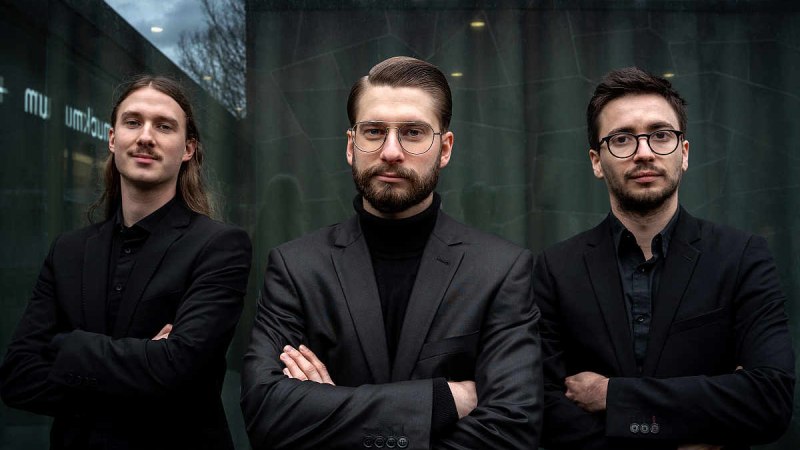 Drei M&auml;nner in schwarzen Anz&uuml;gen stehen mit verschr&auml;nkten Armen vor einer Glaswand. Der Himmel ist bew&ouml;lkt., &copy; JH-Photography