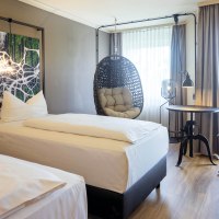 Modernes Hotelzimmer mit zwei Einzelbetten, einem Hängesessel, einem runden Tisch und einem Hocker. Große Fenster mit Vorhängen lassen viel Licht herein., © Seminaris Hotel Bad Boll