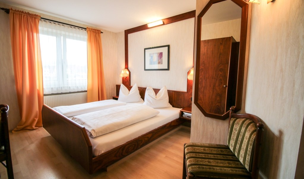 Gemütliches Doppelzimmer mit Holzmöbeln, großem Bett, Spiegel und Fenster mit Vorhängen. Warme Farben und einladende Atmosphäre., © Ulli Kellenbenz