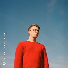 Eine Person in einem roten Pullover steht vor einem klaren blauen Himmel. Der Blick ist nach vorne gerichtet, die Atmosph&auml;re wirkt ruhig und nachdenklich., &copy; links im Bild