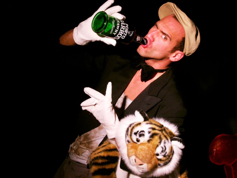 Ein Mann in einem Kostüm trinkt aus einer Flasche und hält einen Plüschtiger. Er trägt weiße Handschuhe und eine Fliege., © Nelly Eichhorn