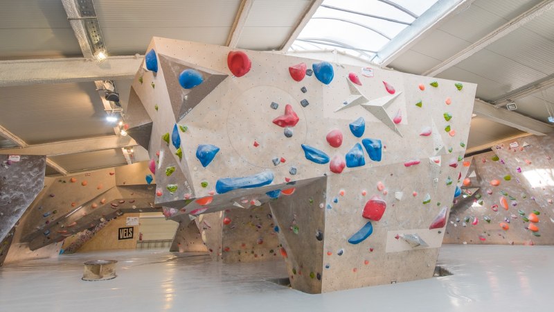 Innenansicht der VELS Boulderhalle mit Kletterwänden und bunten Griffen unter einem gewölbten Dach., © Boulderhalle Vels Innenansicht der VELS Boulderhalle mit Kletterwänden und bunten Griffen unter einem gewölbten Dach., © Boulderhalle Vels