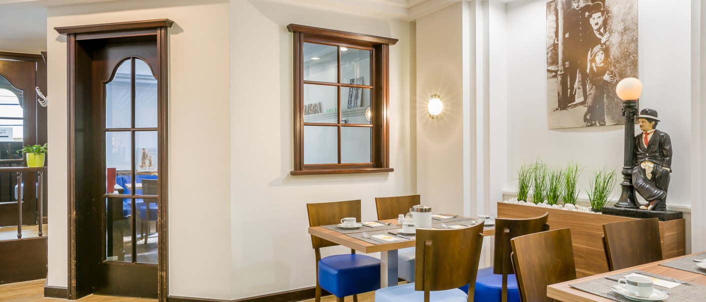 A breakfast room with wooden tables, blue chairs and a statue. A picture hangs on the wall and plants decorate the room., © TOMAS