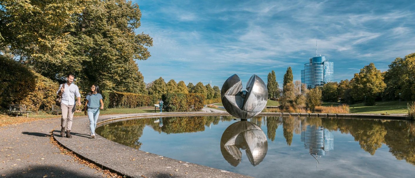 A couple is walking in Wartberg Park along a pond, with a large sculpture and a modern building in the background., © Stuttgart-Marketing GmbH, Sarah Schmid A couple is walking in Wartberg Park along a pond, with a large sculpture and a modern building in the background., © Stuttgart-Marketing GmbH, Sarah Schmid