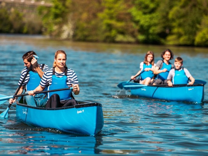 Zwei blaue Kanus mit Menschen paddeln auf einem Fluss. Die Personen tragen gestreifte Shirts und blaue Schwimmwesten. Im Hintergrund ist gr&uuml;ne Natur zu sehen., &copy; Cool-Tours StattReisen