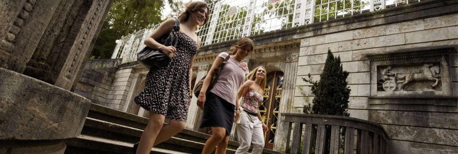 Three women walk down a flight of steps in Stuttgart's Weissenburgpark, past a historic building with a relief and a lattice fence., © Stuttgart-Marketing GmbH Christoph Düpper