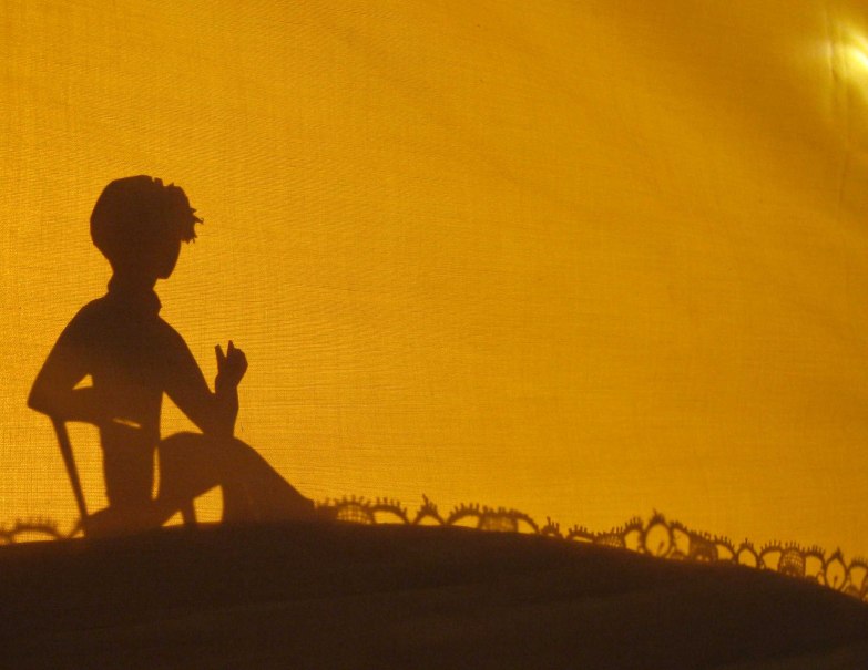 Ein Scherenschnitt zeigt ein Kind, das vor einem gelben Hintergrund sitzt. Die Silhouette ist detailliert und wirkt künstlerisch., © Helen Pavel