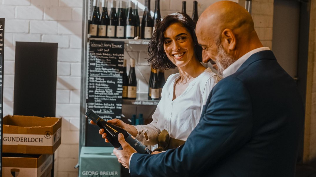 Zwei Personen in einer Weinhandlung lächeln und betrachten gemeinsam eine Weinflasche. Im Hintergrund sind Weinregale und eine Preistafel zu sehen., © SMG, Alwin Maigler Zwei Personen in einer Weinhandlung lächeln und betrachten gemeinsam eine Weinflasche. Im Hintergrund sind Weinregale und eine Preistafel zu sehen., © SMG, Alwin Maigler