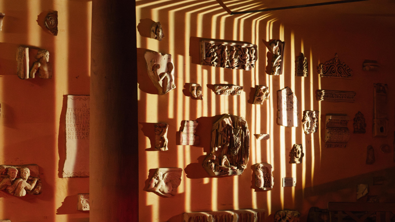 Antike Reliefs und Skulpturen an einer orangefarbenen Wand, beleuchtet von Streifenlicht, das durch eine Säule fällt. Die Szene wirkt warm und atmosphärisch., © Lichtgestalten
