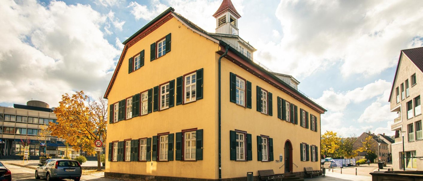 A historic building in Gerlingen with a yellow façade and green shutters, surrounded by modern buildings and autumnal trees., © Stuttgart-Marketing GmbH, Sarah Schmid A historic building in Gerlingen with a yellow façade and green shutters, surrounded by modern buildings and autumnal trees., © Stuttgart-Marketing GmbH, Sarah Schmid