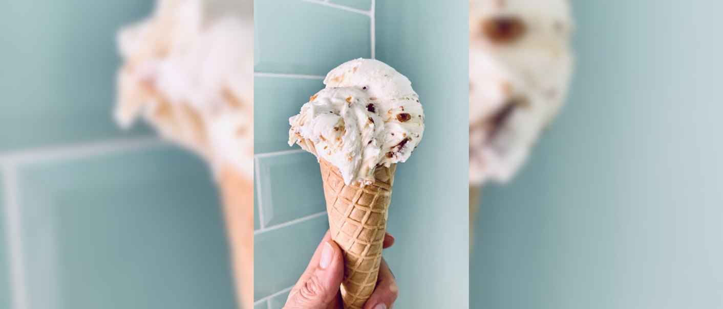 A hand holds an ice cream cone in front of a blue tiled wall. The ice cream has a creamy texture with pieces of nuts., © Zur Schleckerei A hand holds an ice cream cone in front of a blue tiled wall. The ice cream has a creamy texture with pieces of nuts., © Zur Schleckerei