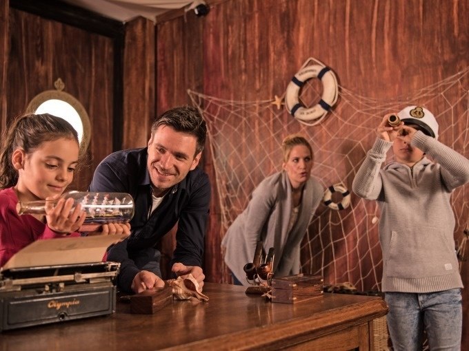 Family in an escape room with a pirate theme. A girl holds a message in a bottle while a boy looks through a telescope. A fishing net in the background., &copy; ExitGames Stuttgart