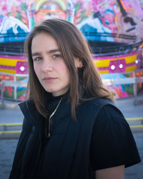 Frau mit braunen Haaren und schwarzer Weste steht vor einem bunten, unscharfen Hintergrund mit leuchtenden Farben und Mustern., &copy; Renitenztheater Stuttgart e.V.
