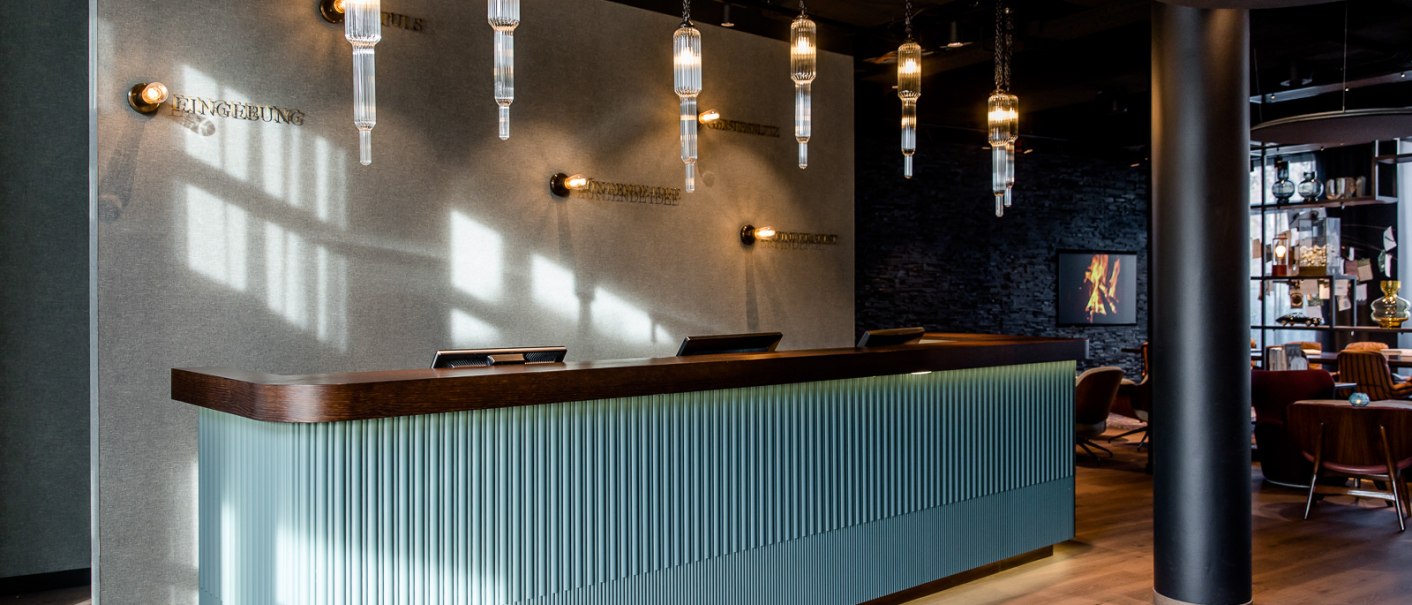 Moderne Rezeption mit hängenden Glasleuchten, Holzdetails und einer strukturierten Front. Im Hintergrund sind dekorative Elemente an der Wand., © TOMAS