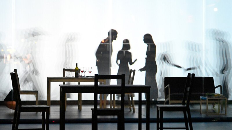 Schatten von Menschen hinter einer beleuchteten Leinwand. Im Vordergrund stehen ein Tisch mit Weingläsern und Stühle. Die Szene wirkt theatralisch., © Württembergische Staatstheater Stuttgart Schatten von Menschen hinter einer beleuchteten Leinwand. Im Vordergrund stehen ein Tisch mit Weingläsern und Stühle. Die Szene wirkt theatralisch., © Württembergische Staatstheater Stuttgart