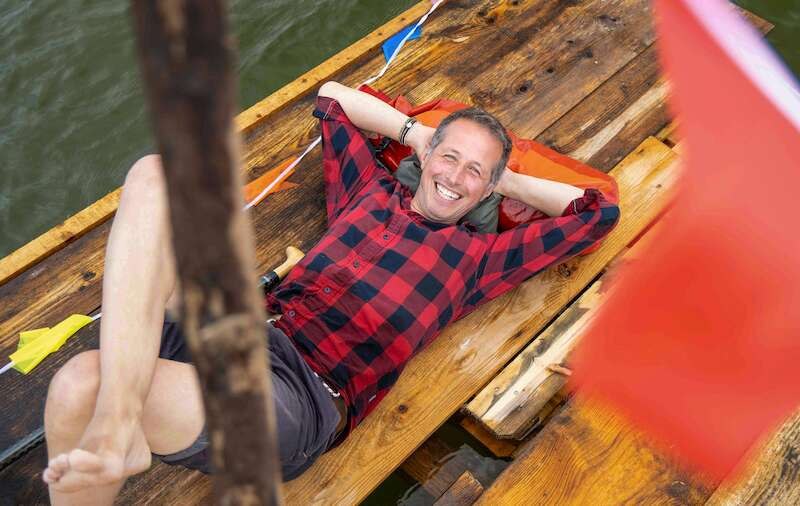 A man in a checked shirt is lying relaxed on a wooden raft on the water. He is smiling and has his arms folded behind his head., &copy; Theaterhaus Stuttgart e.V.