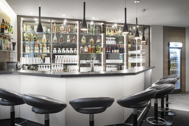 A modern bar with black stools, illuminated shelves filled with bottles and glasses, and stylish pendant lights., &copy; TOMAS