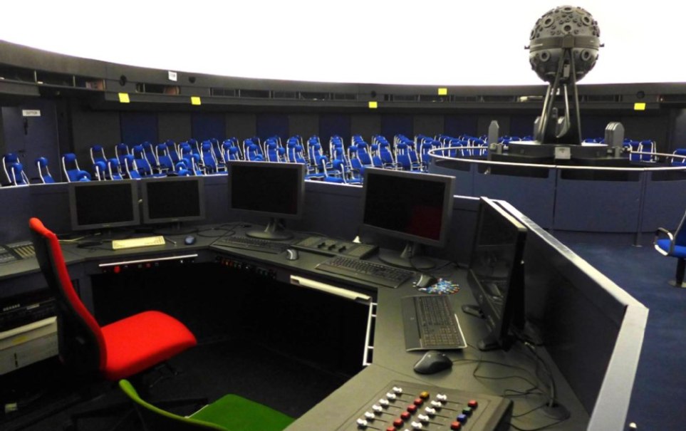 Control panel with screens and buttons in a planetarium. Blue rows of seats and a large projector can be seen in the background., &copy; Planetarium Stuttgart