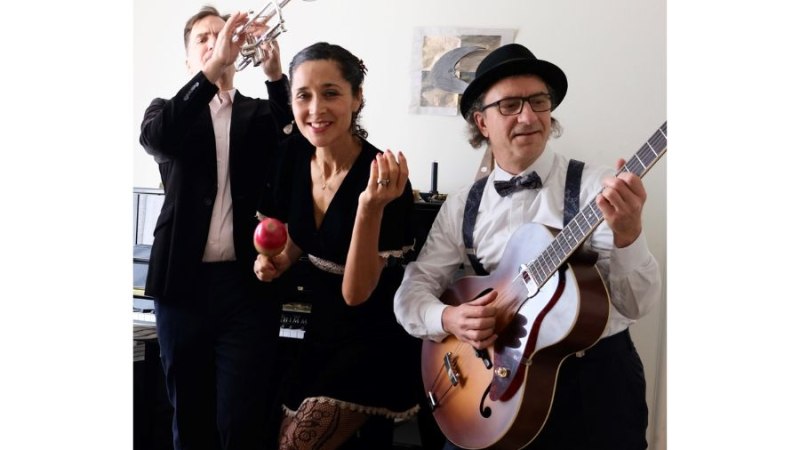 Ein Trio von Musikern in einem Raum: ein Trompeter, eine Frau mit Maraca und ein Gitarrist mit Hut. Sie wirken konzentriert und leidenschaftlich., © PaperMoon