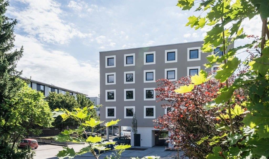 A modern hotel building with square windows, surrounded by green and red plants, under a blue sky., &copy; a2 HOTELS am Quadrium