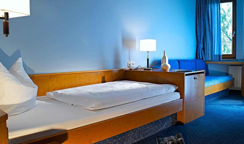 A cozy single room with blue carpet, single bed, sofa and window with blue curtains. A lamp and magazine are visible., © Nödinger Gastro GmbH