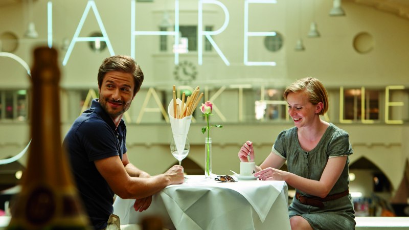 A couple sits smiling at a table in a café. Large letters are visible on the wall in the background., © TMBW, Christoph Düpper