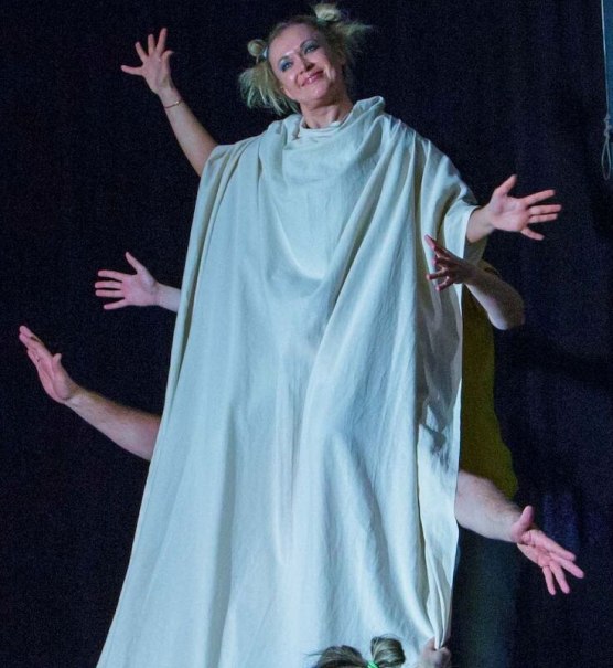 A woman in a white robe with several hands sticking out of the robe, on a stage in front of a dark background., &copy; Theater Atelier