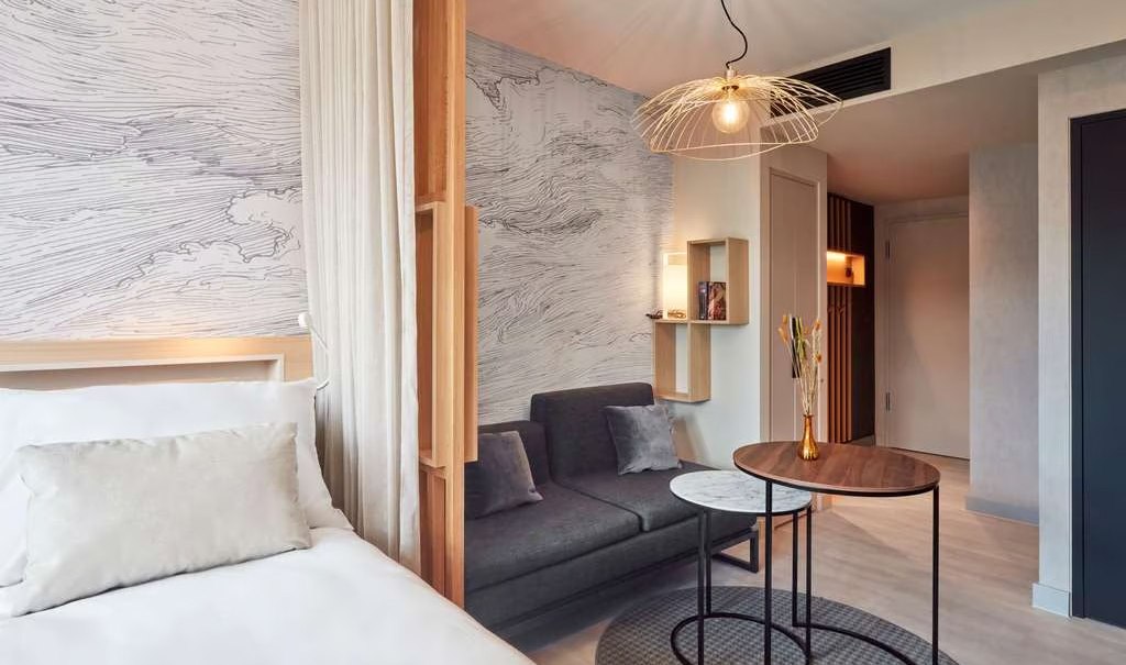 Modernes Studio mit Bett, Sofa, rundem Tisch und stilvoller Lampe. Wand mit abstraktem Muster, helle Farben und minimalistische Einrichtung., © Adagio
