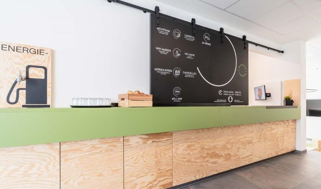 A modern reception area with a green counter, wood paneling, a black information board, a water station, and small plants., &copy; a2 HOTELS am Quadrium