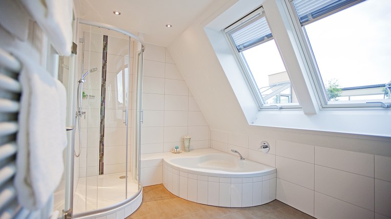 Helles Badezimmer mit Eckbadewanne, runder Duschkabine und großen Dachfenstern. Weiße Fliesen und Handtuchhalter an der Wand., © Hotel Krone Helles Badezimmer mit Eckbadewanne, runder Duschkabine und großen Dachfenstern. Weiße Fliesen und Handtuchhalter an der Wand., © Hotel Krone
