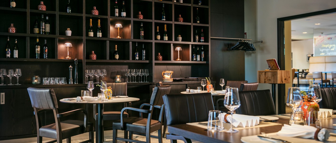 Elegant restaurant with dark wooden furniture, laid tables and a large shelf full of wine bottles and glasses., © ARCOTEL Hotels Elegant restaurant with dark wooden furniture, laid tables and a large shelf full of wine bottles and glasses., © ARCOTEL Hotels