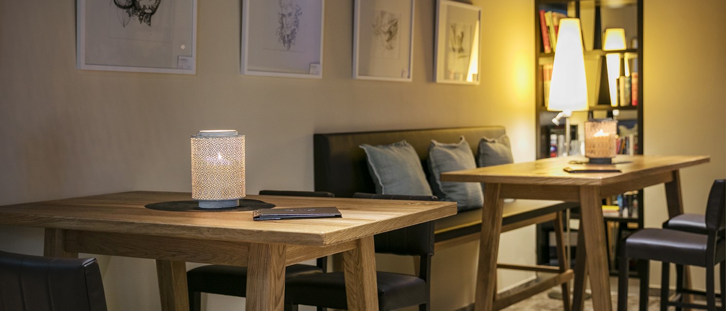 Cozy bar with wooden tables, candlelight and art on the walls. A bookshelf and subtle lighting in the background., © Arcotel Hotels Cozy bar with wooden tables, candlelight and art on the walls. A bookshelf and subtle lighting in the background., © Arcotel Hotels