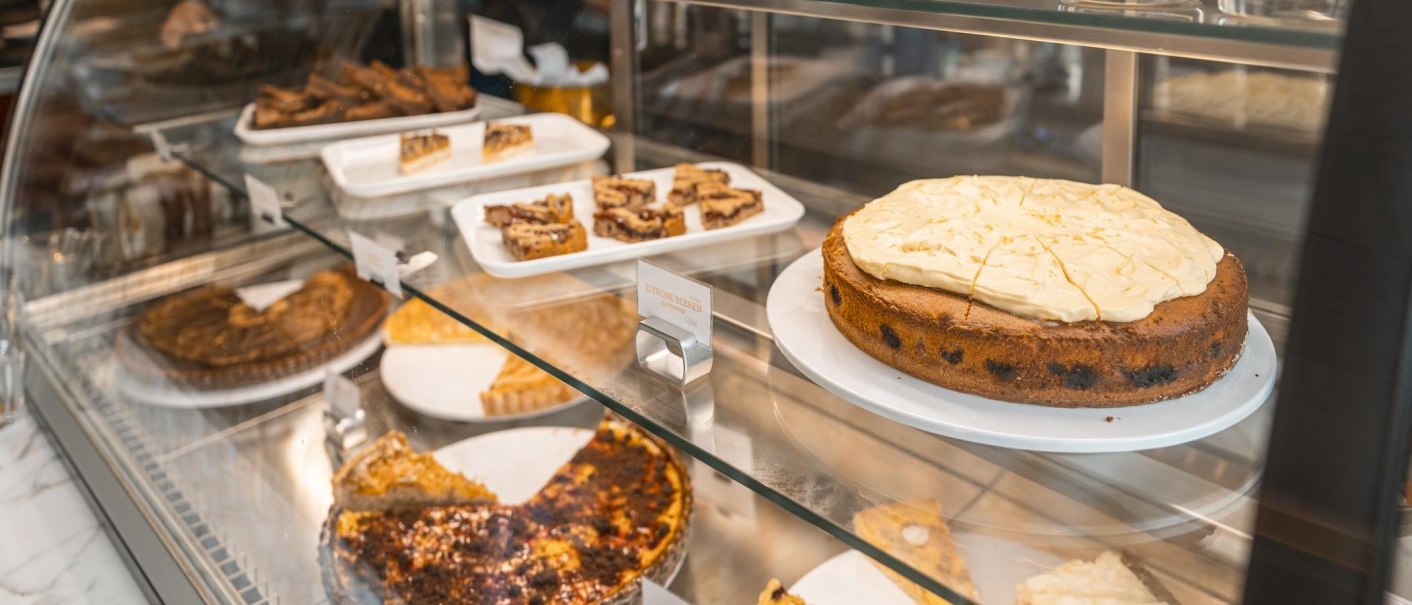 Eine Kuchenvitrine in einem Café zeigt verschiedene Kuchen und Gebäckstücke, darunter ein großer Kuchen mit heller Glasur und kleine Gebäckstücke auf Tabletts., © SMG, Sarah Schmid