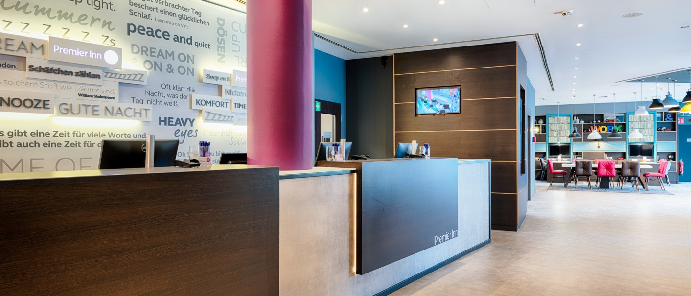 Modern hotel reception from Premier Inn with lettering on the wall and an adjoining seating area with tables and chairs., © Premier Inn Germany Modern hotel reception from Premier Inn with lettering on the wall and an adjoining seating area with tables and chairs., © Premier Inn Germany