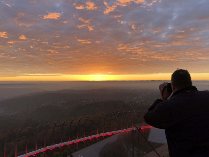 Ein Mann fotografiert den Sonnenuntergang vom SWR Fernsehturm Stuttgart aus. Der Himmel ist orange gef&auml;rbt, und die Landschaft erstreckt sich im Hintergrund., &copy; SWR Media Services GmbH / Fernsehturm Stuttgart