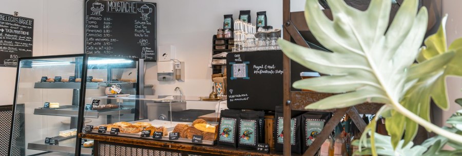 A cozy café with a counter, pastry display and drinks menu. Plants decorate the room and a blackboard displays the menu., © SMG, Sarah Schmid
