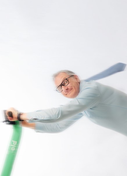 Ein Mann mit Brille f&auml;hrt dynamisch auf einem gr&uuml;nen E-Scooter, seine Krawatte weht im Wind. Der Hintergrund ist wei&szlig;., &copy; Renitenztheater Stuttgart e.V.