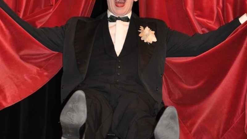 Ein Mann in Frack und Fliege sitzt lachend vor roten Vorhängen und hebt die Beine in die Luft., © Renitenztheater Stuttgart e.V.