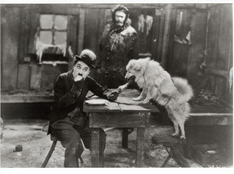 Ein Mann sitzt an einem Tisch und füttert einen Hund, während ein anderer Mann im Hintergrund steht. Die Szene wirkt humorvoll und stammt aus einem alten Film., © Württembergische Staatstheater Stuttgart