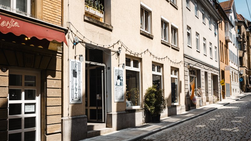 A sunny alleyway with cobblestones, lined with stores such as a second-hand bookshop. Fairy lights hang above the entrances., © SMG Stuttgart Marketing GmbH - Sarah Schmid A sunny alleyway with cobblestones, lined with stores such as a second-hand bookshop. Fairy lights hang above the entrances., © SMG Stuttgart Marketing GmbH - Sarah Schmid