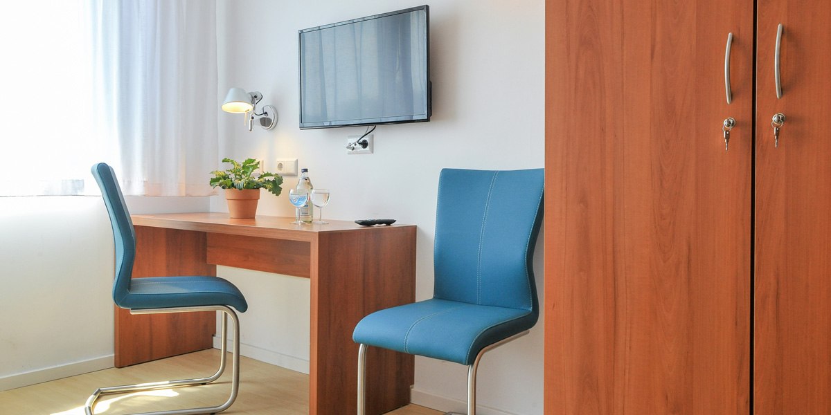 A simple hotel room with a desk, two blue chairs, a TV on the wall, and a closet., &copy; G&auml;stehaus ABZ Stuttgart