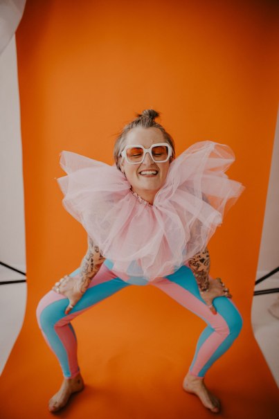 Eine Person in bunter Kleidung und gro&szlig;em T&uuml;llkragen posiert fr&ouml;hlich vor einem orangefarbenen Hintergrund., &copy; Rosenau Kultur e.V.