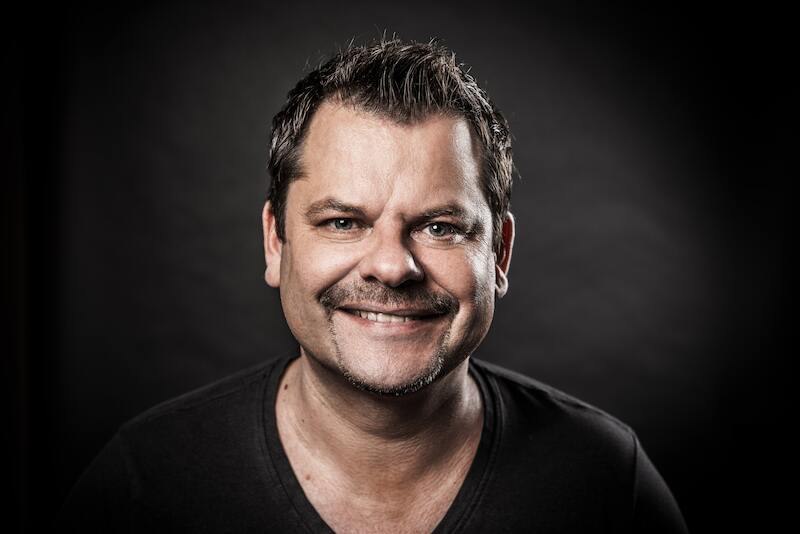 A man with short hair and a beard smiles at the camera. The background is dark, which draws the focus to the face., © SBEntertainment GmbH & Co.KG A man with short hair and a beard smiles at the camera. The background is dark, which draws the focus to the face., © SBEntertainment GmbH & Co.KG
