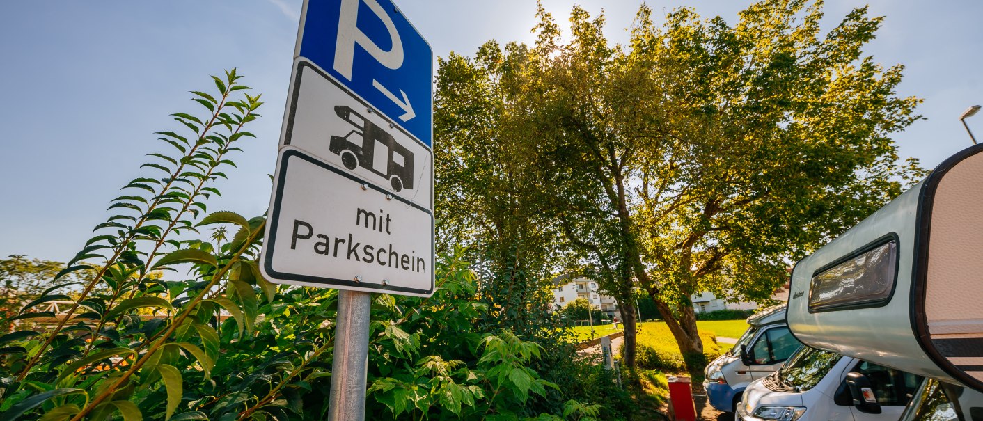 Ein Schild für einen Wohnmobilstellplatz mit Parkschein, umgeben von Bäumen und geparkten Wohnmobilen bei sonnigem Wetter., © Stuttgart-Marketing GmbH, Thomas Niedermüller
