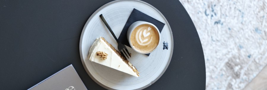 Latte mit Herz-Latte-Art und ein St&uuml;ck Kuchen auf einem Teller auf schwarzem Tisch, daneben eine Karte mit &bdquo;home interior&ldquo;., &copy; Taraba