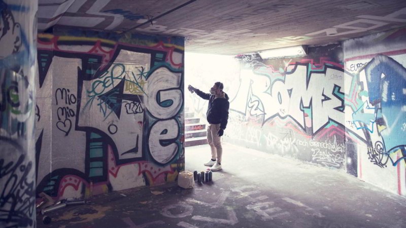 Eine Person sprüht Graffiti in einem Tunnel. Die Wände sind mit bunten Graffitis bedeckt. Auf dem Boden stehen Farbdosen., © hoponopo GmbH